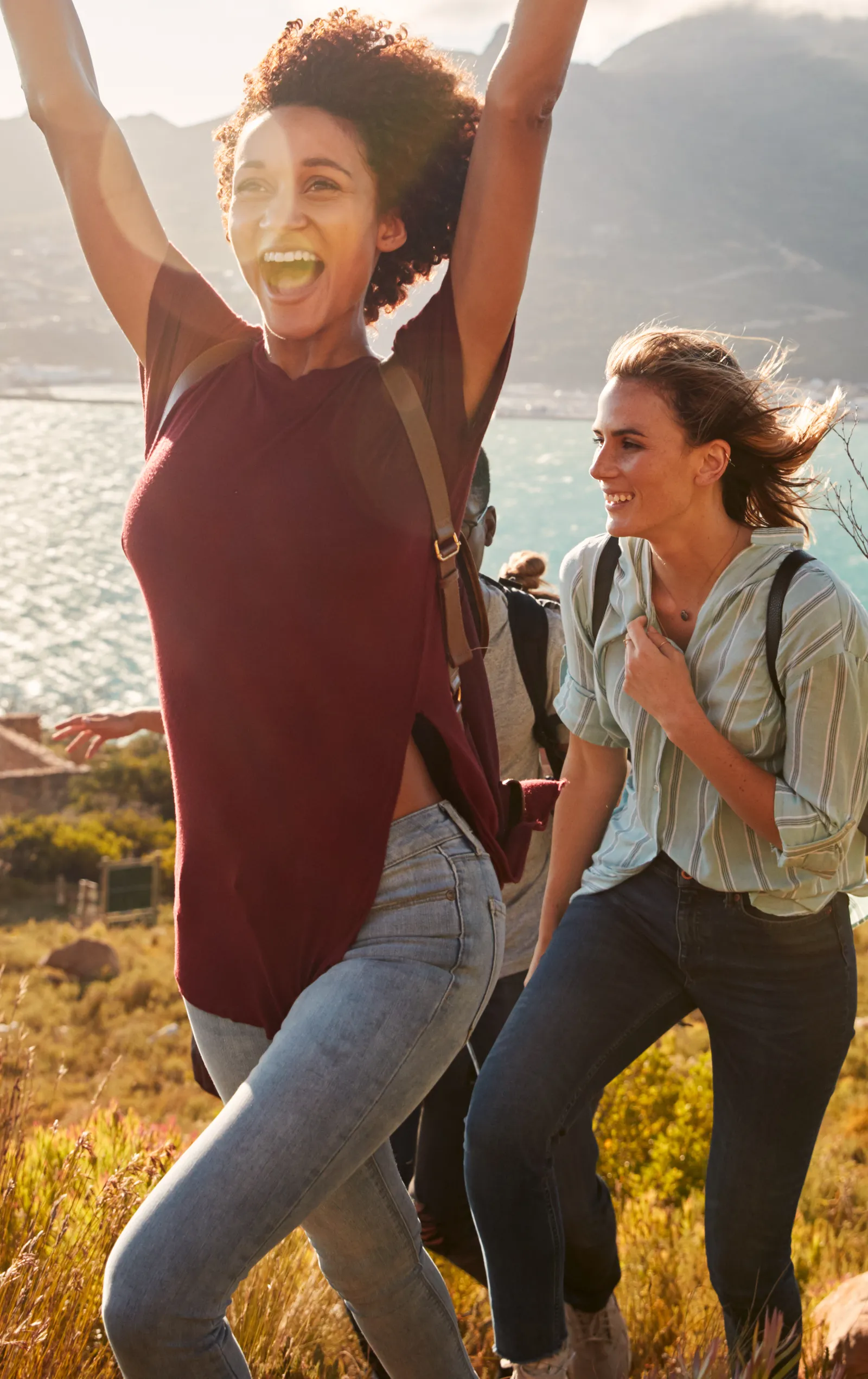 Women hiking a coastal ridge in late afternoon sun, one throwing both arms up in celebration, a turquoise bay and mountains behind them